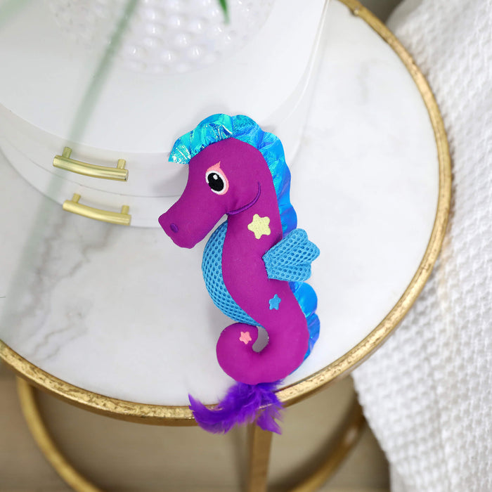 The Quirky Kitty Sea Horse Kicker, an ocean-themed cat kicker toy with catnip, featuring a purple body, blue mane and fins, star accents, and a feather tail by Quirky Kitty, rests on a round white and gold table.