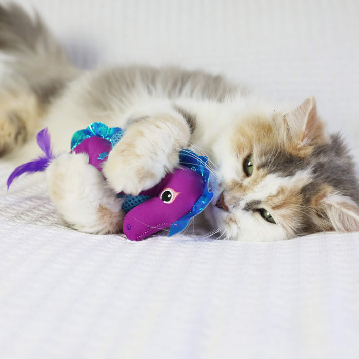A fluffy cat lounges on a white surface, playfully hugging and biting the Quirky Kitty Sea Horse Kicker, an ocean-themed cat kicker toy with catnip from Quirky Kitty.