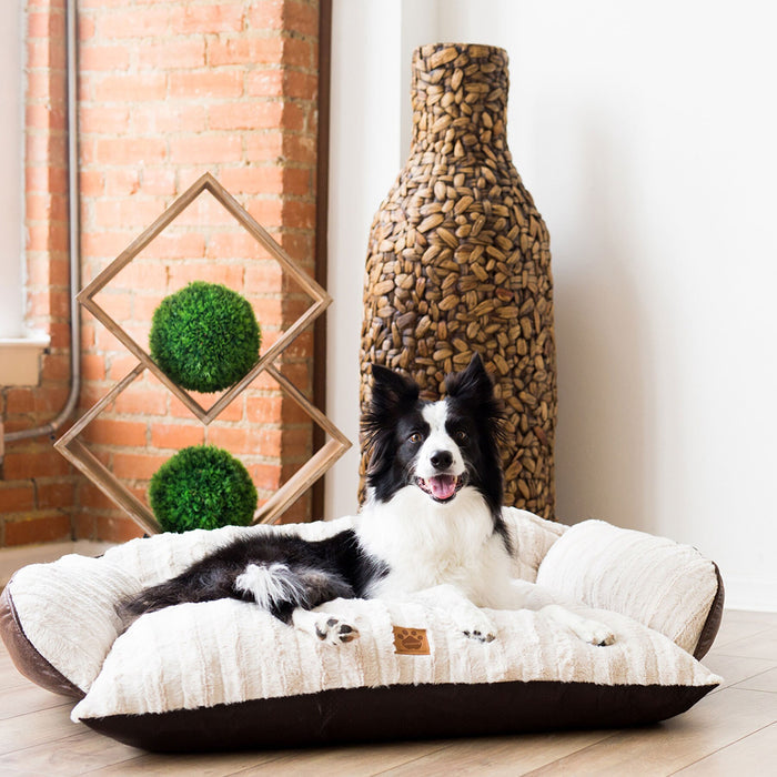 A black and white dog sits happily on a plush white SnooZZy Rustic Luxury Comfy Couch Pet Bed by Snoozzy in a room with a brick wall. A textured vase and geometric wooden sculpture with green spheres enhance the background. The bed is machine washable, blending relaxation with practicality.