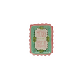 A small rectangular ceramic dish with a scalloped pink border and a turquoise center, featuring the words “MERRY and BRIGHT” in pink script surrounded by green leafy illustrations. The dish is photographed against a plain, textured off-white wall.