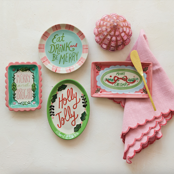 A styled flat-lay of a holiday table setting featuring a pink and white striped plate that reads 'Eat DRINK and BE MERRY', a green oval plate reading 'Holly Jolly', a pink rectangular dish with 'MERRY CHRISTMAS', a pink napkin, a yellow spoon, and a pink textured ornament on a textured off-white background.