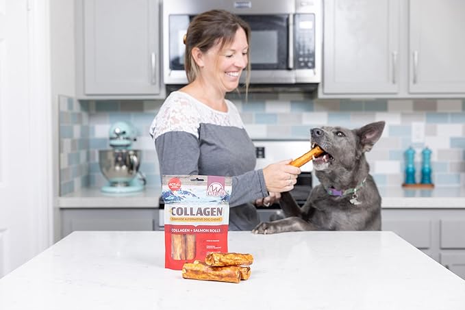 A woman in the kitchen smiles as she holds a Wild Eats Salmon Flavored Collagen Retriever Adult Roll for her happy gray dog on its hind legs. A bag of Wild Eats rolls and digestible dog treats are on the counter in front.